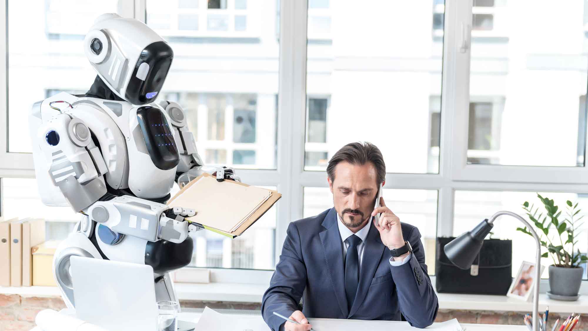 Concentrated serious businessman is holding pencil and talking on smartphone while his assistant robot is passing him envelopes